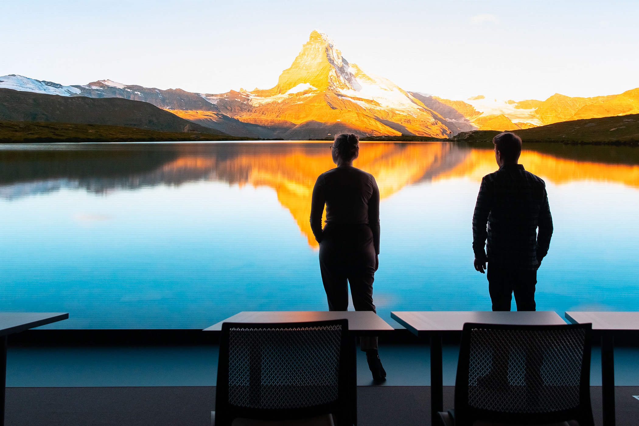 Man and woman in the forefront looking at a screen with an image of a lake by the mountains on a clear daybackground