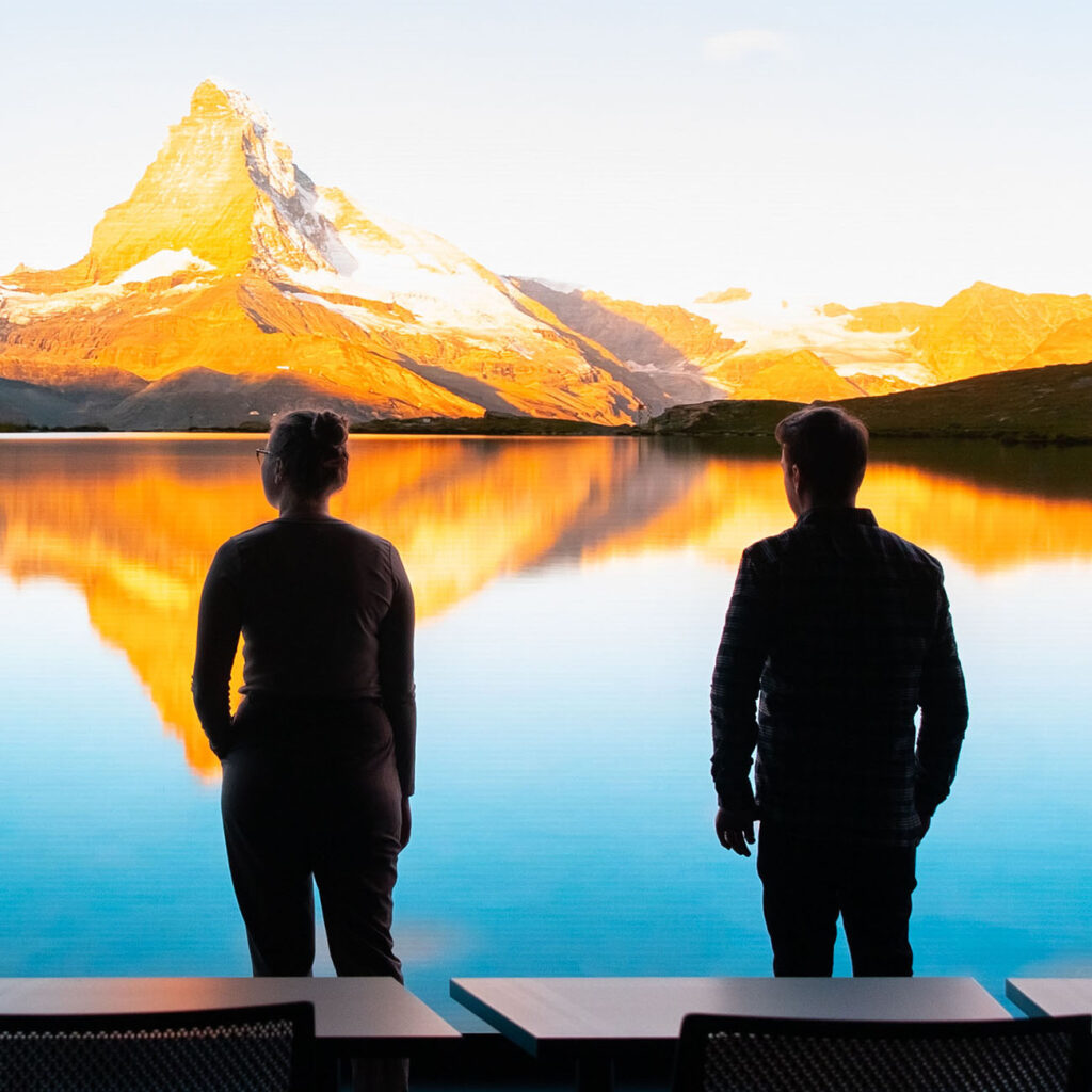 Man and woman in the forefront looking at a screen with an image of a lake by the mountains on a clear daybackground
